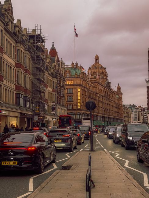 A busy street in the Harrods area of Knightsbridge during daytime, featuring a lineup of parked cars along the curb and moving vehicles including a red double-decker bus. Tall historic buildings with 